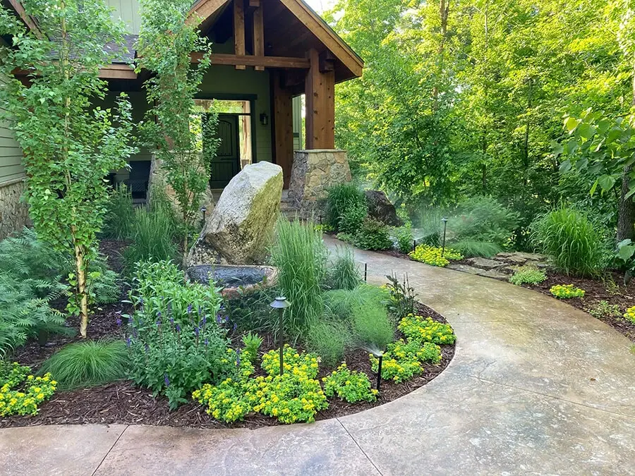 Sprinklers watering flowers and ornamental grasses beside a stone path and timber-framed entrance